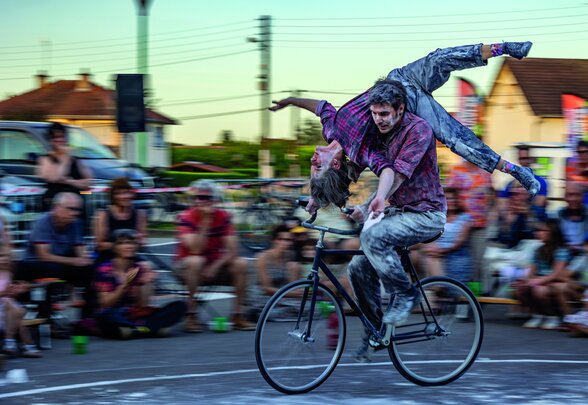 Un acrobate pédalant porte son partenaire renversé et à l'horizontale sur ses épaules, entouré d'un public assis en plein air. - Agrandir l'image, fenêtre modale