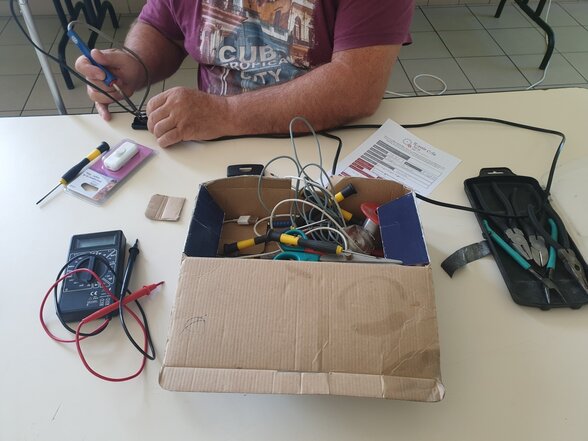 Bénévole réparant un appareil électronique, entouré d'outils : multimètre, tournevis, pinces et câbles sur une table - Agrandir l'image, fenêtre modale