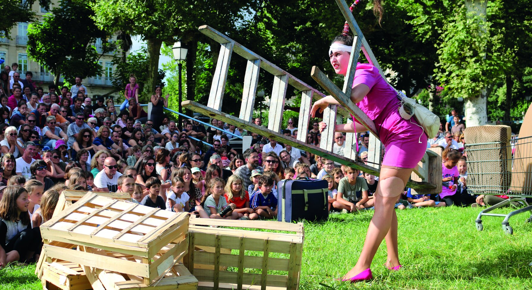 Comédienne en robe rose portant une échelle au-dessus de caisses en bois, jouant en plein air devant un large public assis sur l'herbe