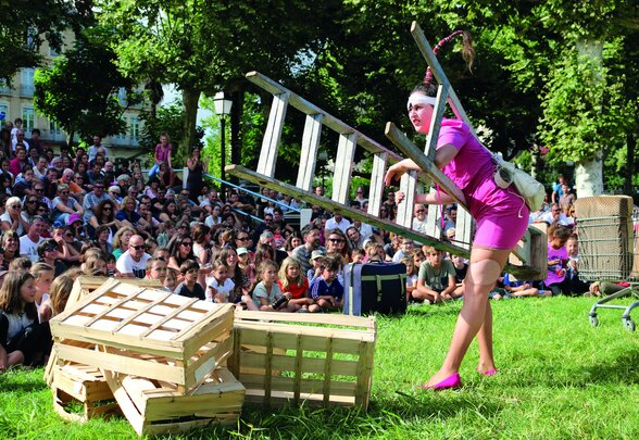 Comédienne en robe rose portant une échelle au-dessus de caisses en bois, jouant en plein air devant un large public assis sur l'herbe - Agrandir l'image, fenêtre modale