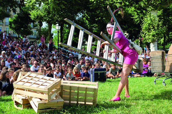 Comédienne en robe rose portant une échelle au-dessus de caisses en bois, jouant en plein air devant un large public assis sur l'herbe - Agrandir l'image, fenêtre modale