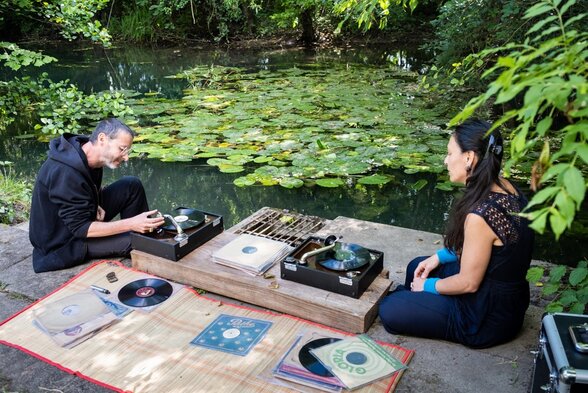 Deux artistes assis face à face autour de platines vinyles au bord d'un étang couvert de nénuphars - Agrandir l'image, fenêtre modale