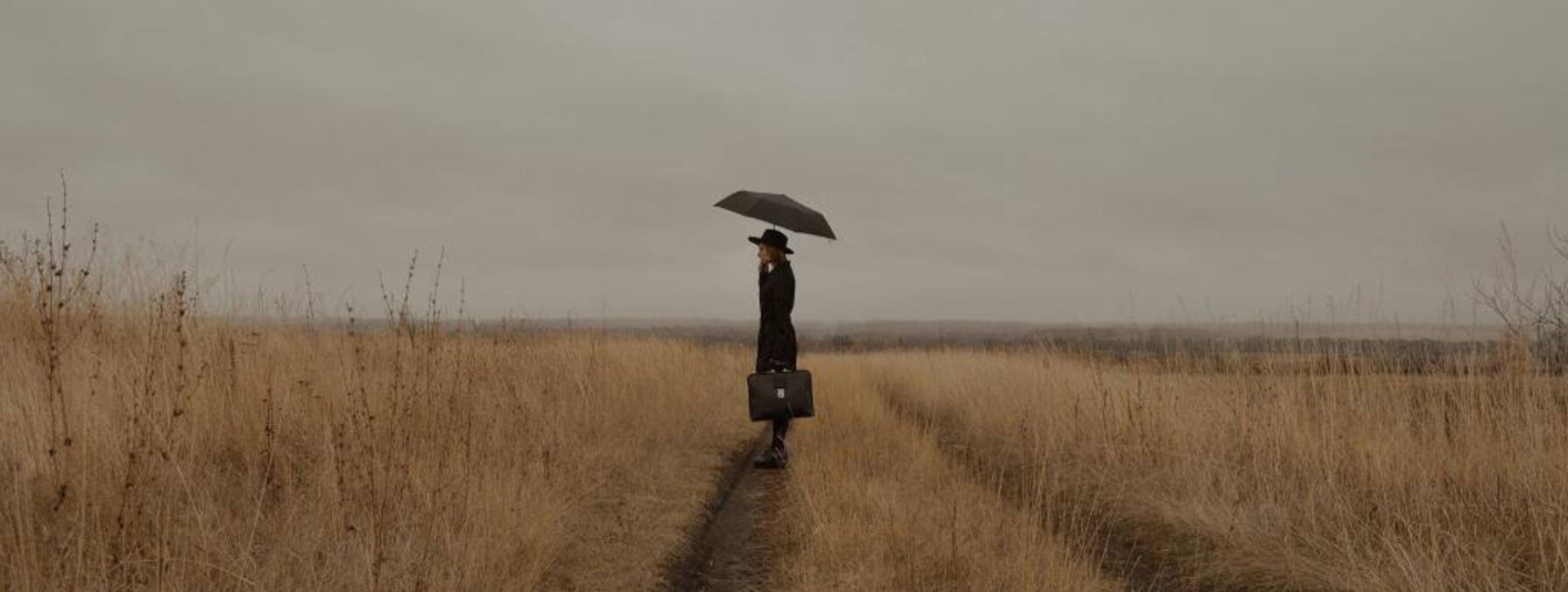une femme dans un champ. Elle s'abrite sous un parapluie et tient une sacoche à la main.
