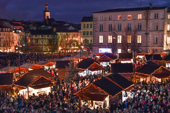 Chalets en bois garnis de guirlandes lumineuses et foule dense place du Donjon en soirée - Agrandir l'image, fenêtre modale