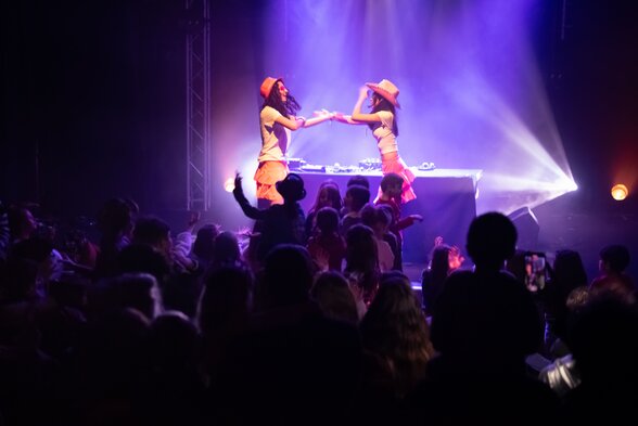 Deux danseuses en costumes colorés se produisent sur scène devant une foule d'enfants sous des jeux de lumières violettes - Agrandir l'image, fenêtre modale