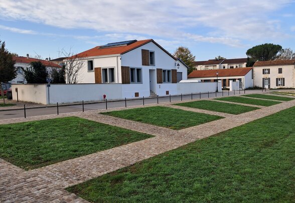 Extérieur rénové du site : bâtiment blanc aux volets bois et toiture tuiles, précédé d'une pelouse structurée par des allées pavées - Agrandir l'image, fenêtre modale