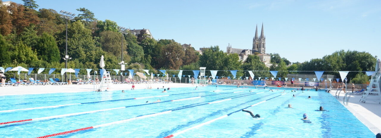 Bassin extérieur de natation avec des nageurs dans les couloirs, l'église Saint-André de Niort visible en arrière-plan par temps ensoleillé