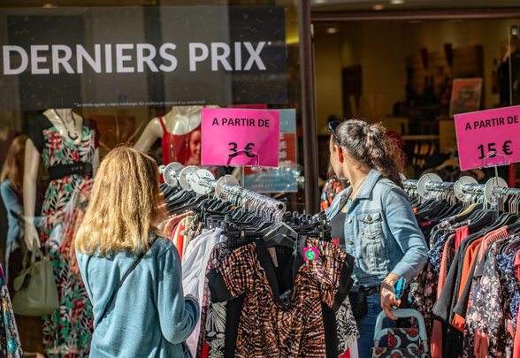 Deux visiteurs parcourent des portants de vêtements, étiquettes indiquant des articles à partir de 3 € et 15 € - Agrandir l'image, fenêtre modale