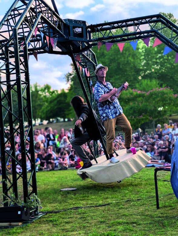 Acrobate en chemise fleurie en équilibre sur une structure métallique en hauteur, face à un large public assis sur l'herbe. - Agrandir l'image, fenêtre modale