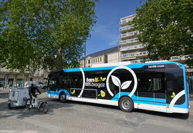 Bus articulé roulant au biogaz portant le marquage « TanLib 100 % biogaz », stationné sur la place de la Brèche