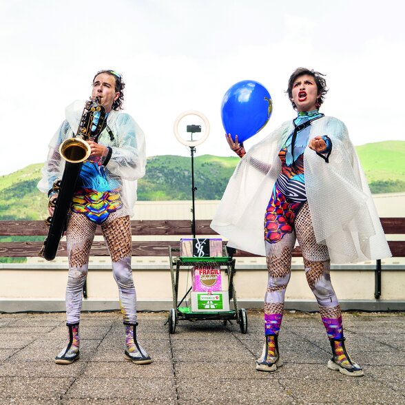 Deux artistes en costumes bariolés et capes plastique, l'un au saxophone, l'autre tenant un ballon bleu, devant un ring light de scène - Agrandir l'image, fenêtre modale