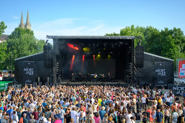 Concert du Niort Jazz Festival en plein air, scène illuminée face à une foule en liesse, clochers de la ville en arrière-plan - Agrandir l'image, fenêtre modale