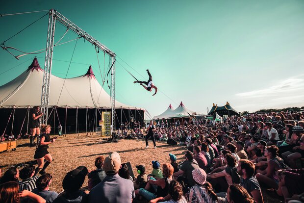 Un acrobate en plein saut périlleux aérien suspendu à une structure métallique, devant une foule nombreuse assise sous des chapiteaux de festival