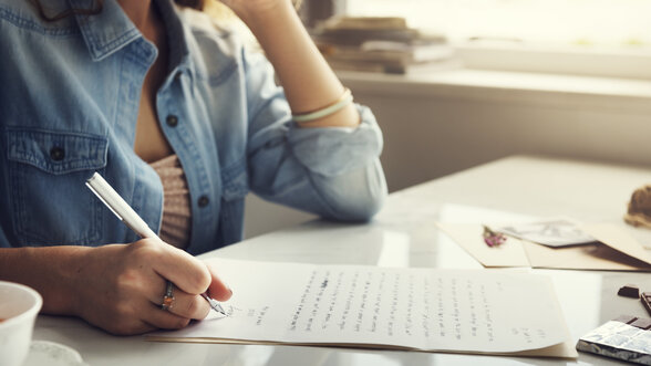 Une personne écrit à la plume sur des feuilles manuscrites posées sur une table, dans une atmosphère lumineuse et apaisante. - Agrandir l'image, fenêtre modale