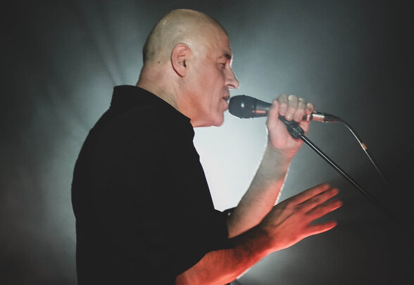 Dominique A en concert, chantant micro en main, guitare en bandoulière, sous un éclairage de scène contrasté - Agrandir l'image, fenêtre modale