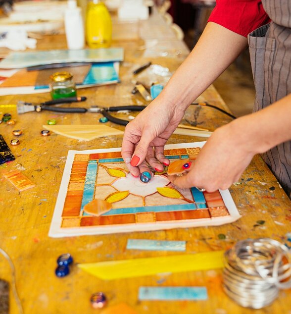 Mains assemblant des pièces de verre coloré sur une composition florale aux tons chauds, outils et fragments de verre dispersés sur un établi d'atelier. - Agrandir l'image, fenêtre modale
