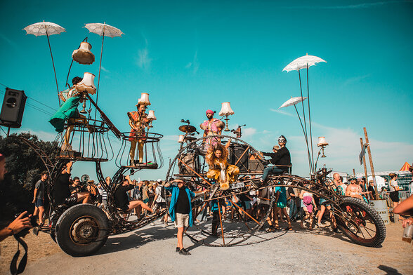 Engin de métal sur roues géantes, artistes en costumes colorés perchés à différents niveaux, parasols blancs et lampes, entouré d'un public - Agrandir l'image, fenêtre modale