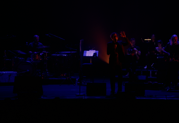Jarvis Cocker en concert, bras levé face au public, accompagné d'un grand orchestre baigné de lumières violettes - Agrandir l'image, fenêtre modale