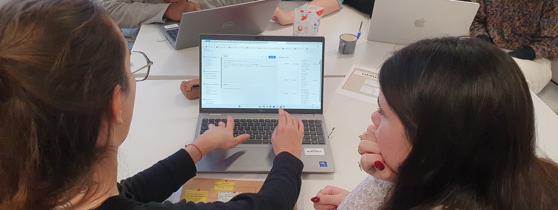 Femmes réunies autour d'ordinateurs portables lors d'un atelier collaboratif, l'une désignant une interface à sa voisine