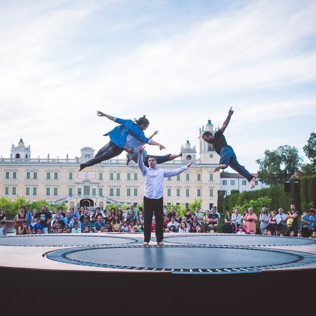 Deux artistes bondissent haut dans les airs au-dessus du plateau circulaire, devant une foule nombreuse et un palais en plein air