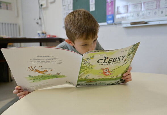 Enfant lisant l'album « Clebsy, chien de la jungle », assis à une table dans une salle d'activités - Agrandir l'image, fenêtre modale