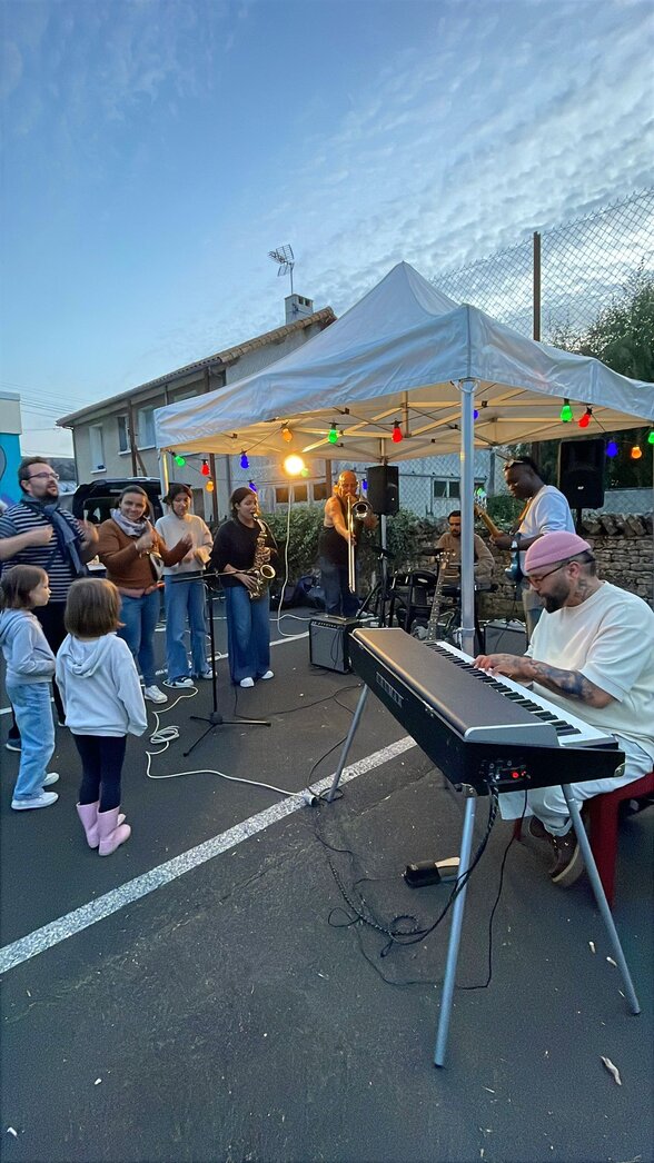 Un groupe de musiciens – clavier, saxophone, trombone – joue en plein air sous un barnum coloré, entouré de familles. - Agrandir l'image, fenêtre modale