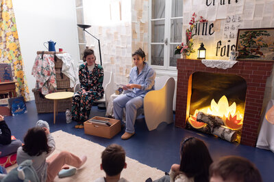 Deux animatrices en pyjama lisent à des enfants assis au sol, dans un décor de cheminée et murs tapissés de pages de livres.