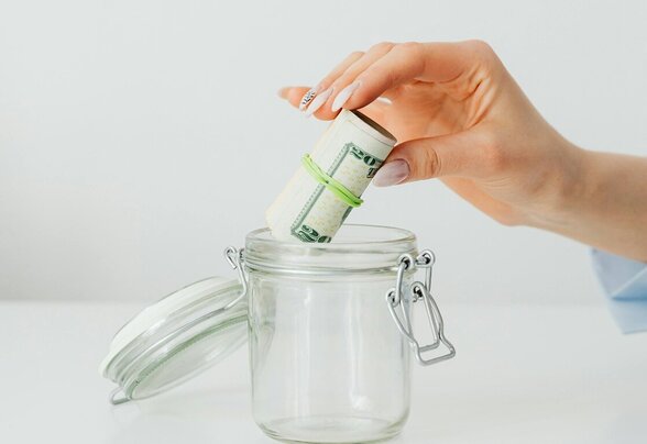 Une main dépose un rouleau de billets de banque dans un bocal en verre, évoquant l'épargne ou la gestion monétaire - Agrandir l'image, fenêtre modale