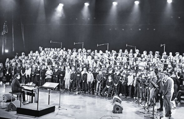 Quelque 400 élèves choristes en formation sur scène sous les projecteurs, accompagnés d'un pianiste - Agrandir l'image, fenêtre modale