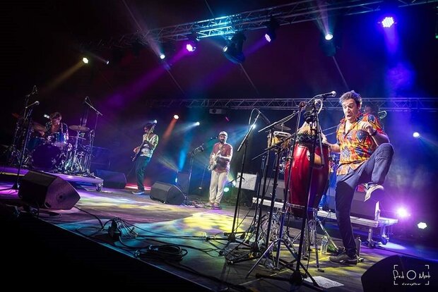Concert sur scène : percussionniste au premier plan sur congas, guitaristes et batteur en arrière-plan sous des lumières colorées