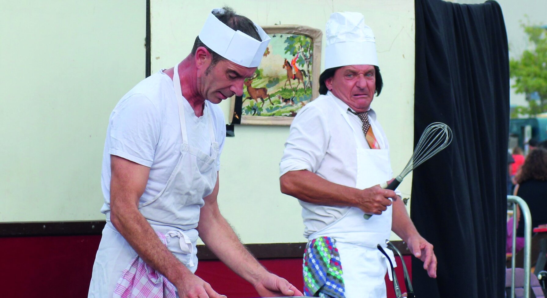 Deux artistes en costume de cuisinier jouent une scène comique en plein air, devant un décor de roulotte et un public