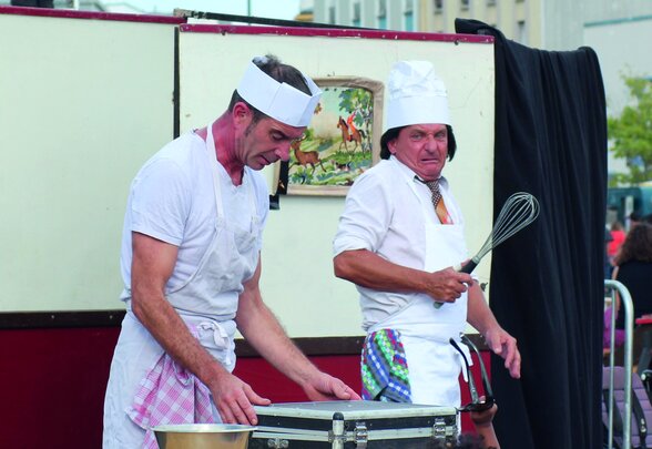 Deux artistes en costume de cuisinier jouent une scène comique en plein air, devant un décor de roulotte et un public - Agrandir l'image, fenêtre modale