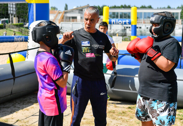 Un entraîneur de boxe donnant des conseils à deux enfants équipés de casques et gants, à Niort Plage - Agrandir l'image, fenêtre modale