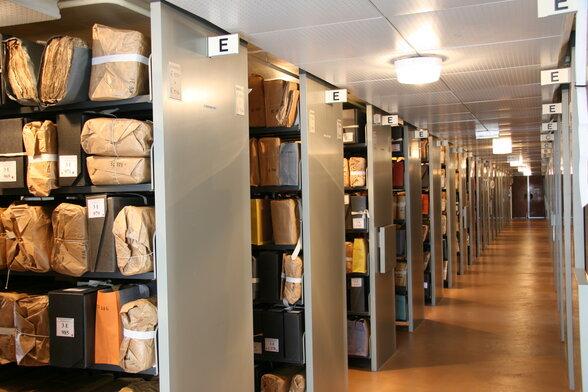 Longue allée de rayonnages métalliques chargés de liasses ficelées et boîtes d'archives dans une salle de conservation. - Agrandir l'image, fenêtre modale