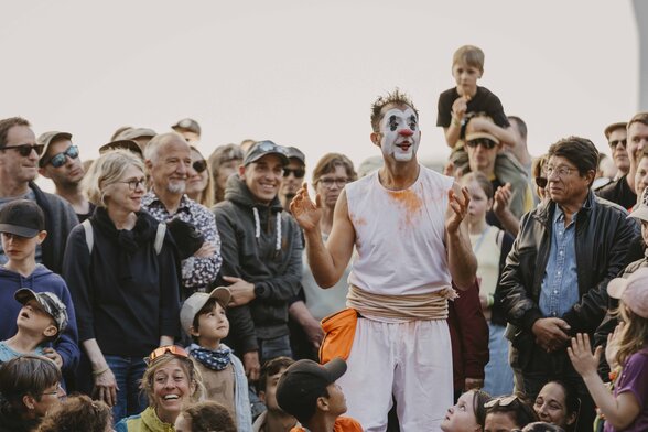 Tuga en costume blanc et maquillage de clown, bras levés, entouré d'un large public souriant en plein air - Agrandir l'image, fenêtre modale