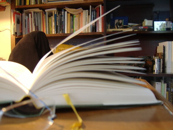 Livre ouvert sur un bureau, pages éventées, devant une bibliothèque garnie d'ouvrages en arrière-plan - Agrandir l'image, fenêtre modale