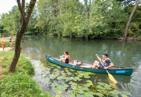 Deux personnes pagayent sur une rivière calme bordée de nénuphars, tandis que des visiteurs se regroupent sur la berge verdoyante - Agrandir l'image, fenêtre modale