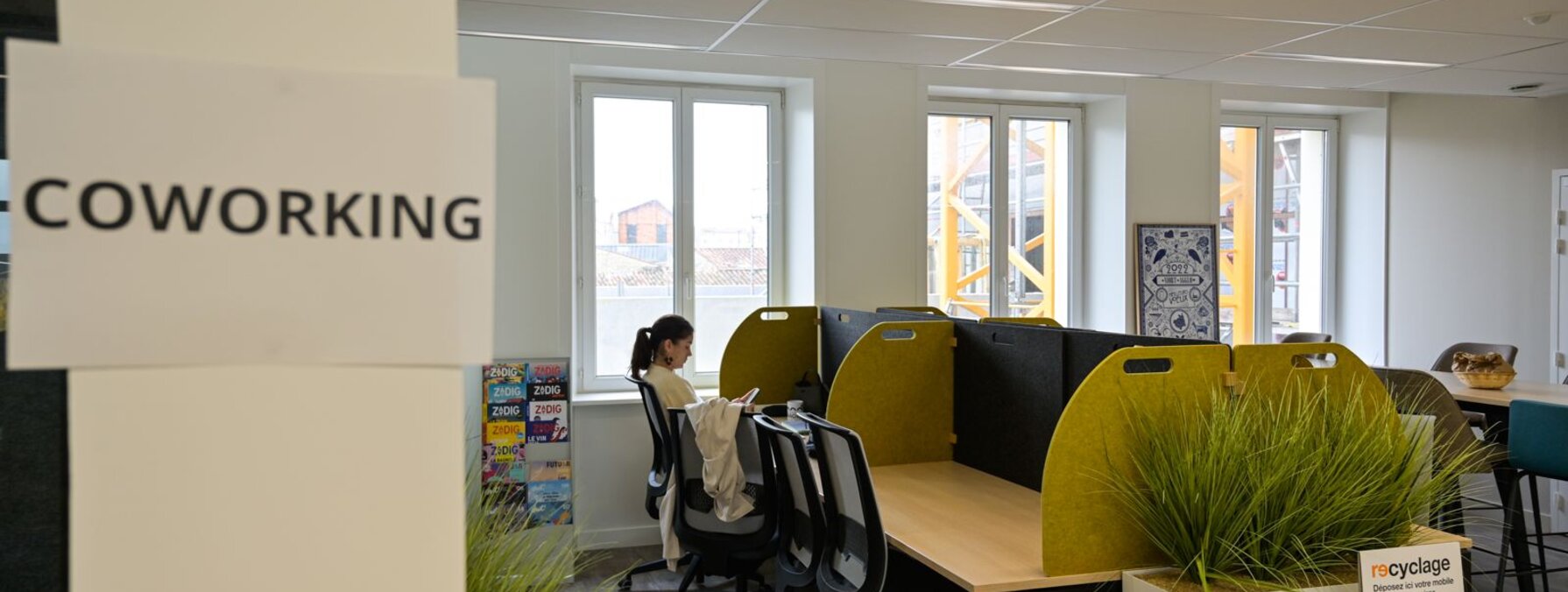 Salle de coworking réaménagée avec postes de travail équipés de cloisons acoustiques, grue de chantier visible par les fenêtres