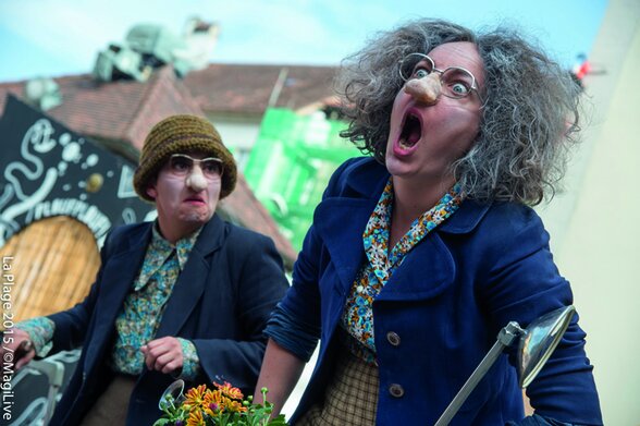 Les deux Germaine en costume scénique : masques à gros nez, perruques grises et vêtements fleuris, jouant en extérieur avec expressivité. - Agrandir l'image, fenêtre modale