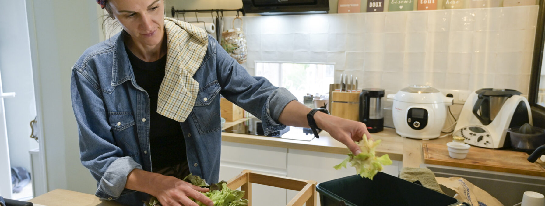 Une personne dépose des feuilles de salade dans un bac de tri des biodéchets, entourée de légumes frais sur un plan de travail.
