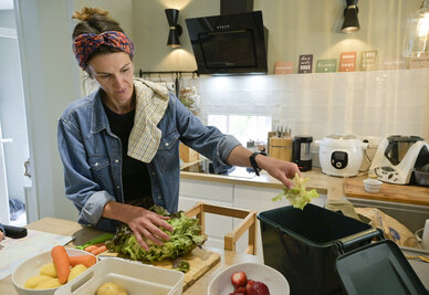 Une personne dépose des feuilles de salade dans un bac de tri des biodéchets, entourée de légumes frais sur un plan de travail.