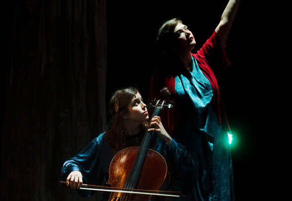 Dom La Nena, violoncelliste assise, et Rosemary Standley, chanteuse bras levé, lors d'une mise en scène théâtrale en forêt. - Agrandir l'image, fenêtre modale