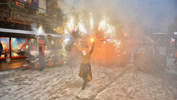 Artiste en costume de scène jonglant avec des feux d'artifice dans une rue enneigée, petit train décoré en arrière-plan - Agrandir l'image, fenêtre modale