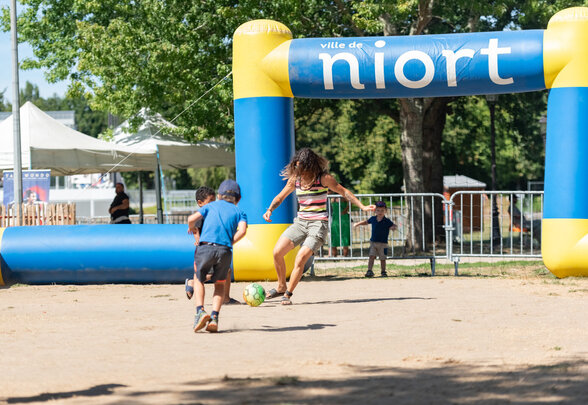 Un adulte et deux enfants jouent au football en plein air sous l'arche gonflable bleue et jaune Ville de Niort. - Agrandir l'image, fenêtre modale