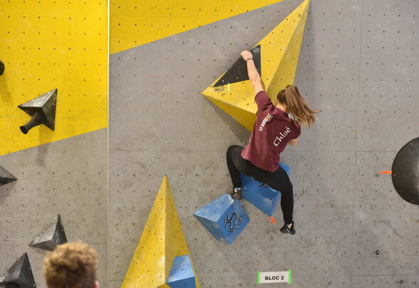 Grimpeuse en action sur le mur de blocs, agrippant une prise géométrique jaune au niveau du bloc 2 - Agrandir l'image, fenêtre modale