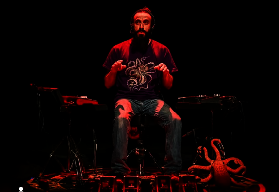Kloctopus sur scène, entouré de percussions aux pieds et claviers, sous un éclairage rouge, avec une pieuvre décorative - Agrandir l'image, fenêtre modale
