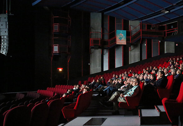 Salle de projection obscure aux rangées de fauteuils rouges, partiellement occupée par des spectateurs attentifs. - Agrandir l'image, fenêtre modale