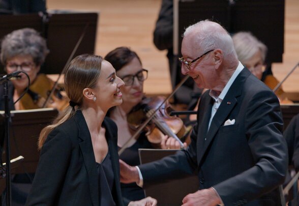 Une soliste et le chef d'orchestre se saluent chaleureusement sur scène, entourés des musiciens - Agrandir l'image, fenêtre modale