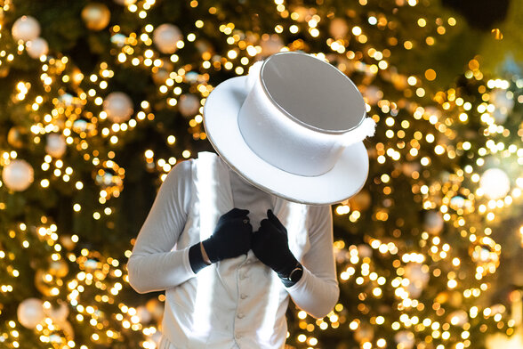Personnage en costume blanc intégral coiffé d'un grand chapeau haut-de-forme, devant un sapin illuminé de lumières dorées - Agrandir l'image, fenêtre modale