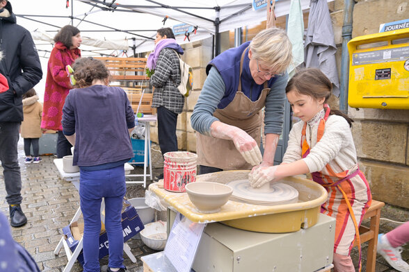 Une animatrice guidant un enfant sur un tour de poterie en plein air, tous deux les mains couvertes d'argile - Agrandir l'image, fenêtre modale